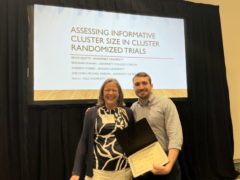 Two people stand in front of screen with presentation title projected on it. One of them holds an open certificate folder.